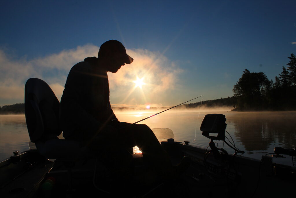 Man i keps sitter och fiskar i en båt. Det ligger dimma över sjön där solen går upp och ger motljus över fiskaren. Himmeln är blå och det är skog runt sjön.